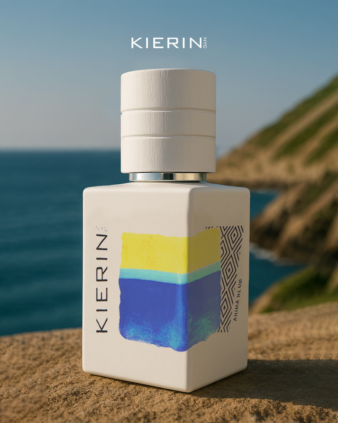 A white bottle of KIERIN NYC perfume with a colorful abstract label sits on a stone surface near the ocean and a grassy cliff.