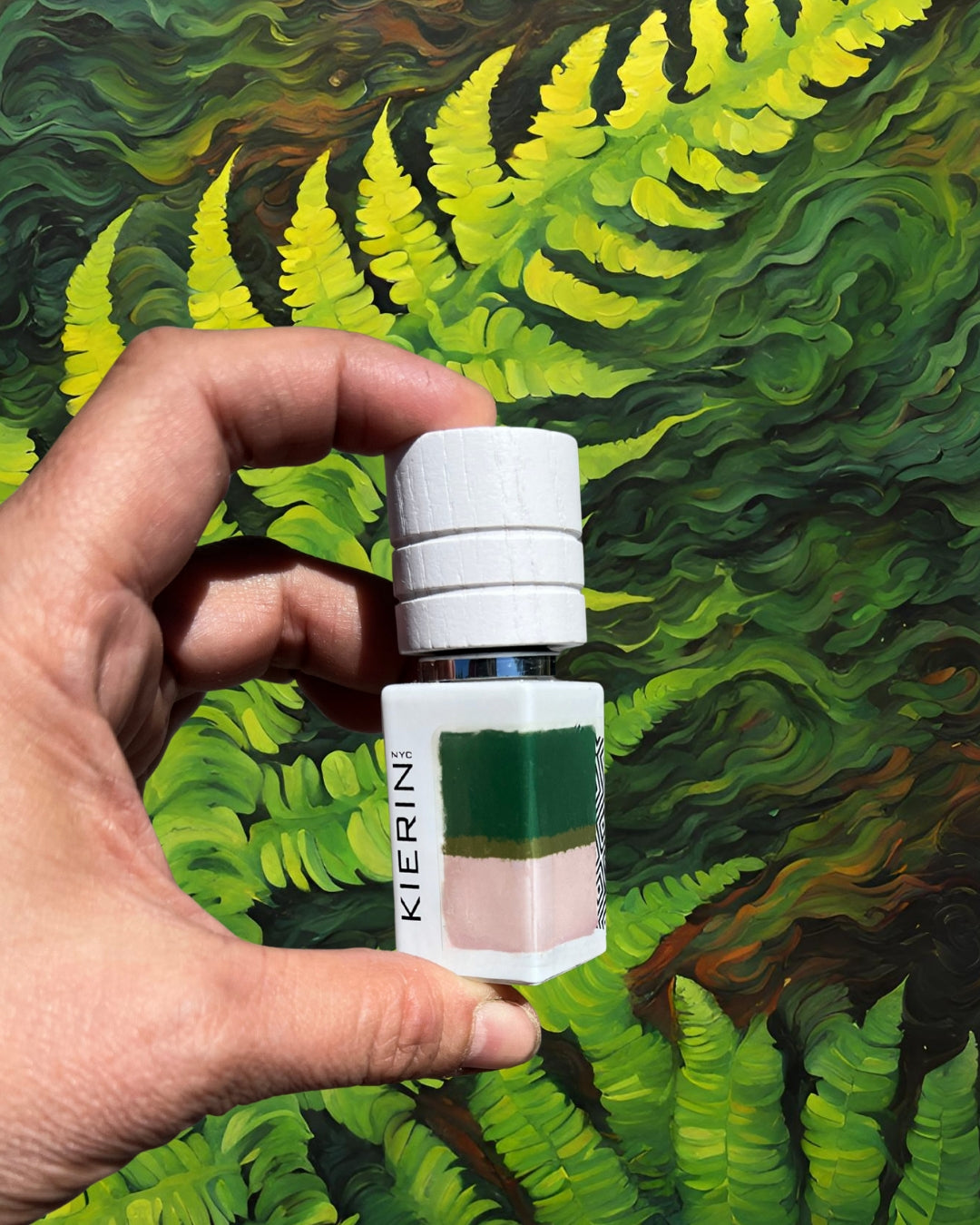 A hand holds a small glass bottle labeled "Kierin NYC" against a vibrant background of lush green ferns, conveying freshness and nature.