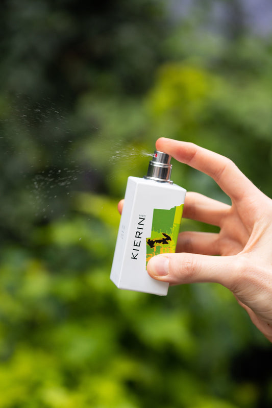 A hand sprays a white rectangular perfume bottle labeled "Kierin" amidst a blurred green leafy background, conveying a fresh, natural vibe.