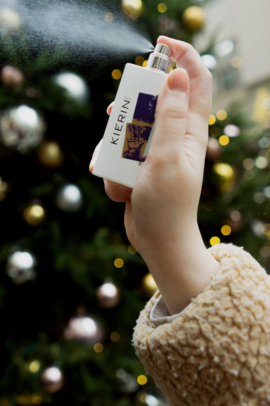 A person sprays perfume from a KIERIN bottle, with a decorated Christmas tree in the blurred background.