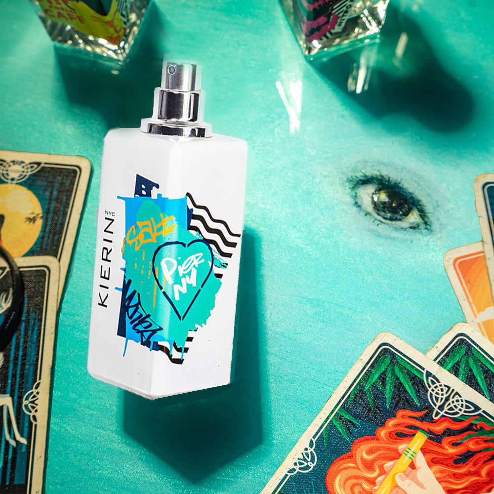 A white perfume bottle with graffiti design stands on a turquoise surface, surrounded by colorful tarot cards and a painted eye, creating a mystical vibe.