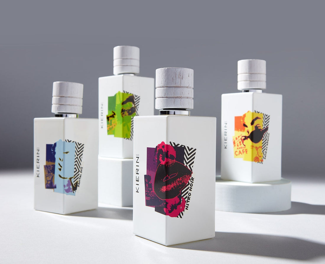 Four elegant perfume bottles with bold, abstract labels in vibrant colors on sleek, white surfaces are displayed on a gradient gray background.