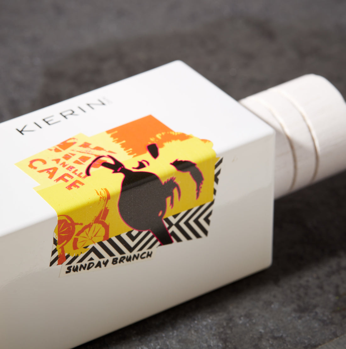 Minimalist white box with colorful urban-themed sticker featuring a stylized cafe scene, cyclist, and text "Sunday Brunch." Energetic and modern design.